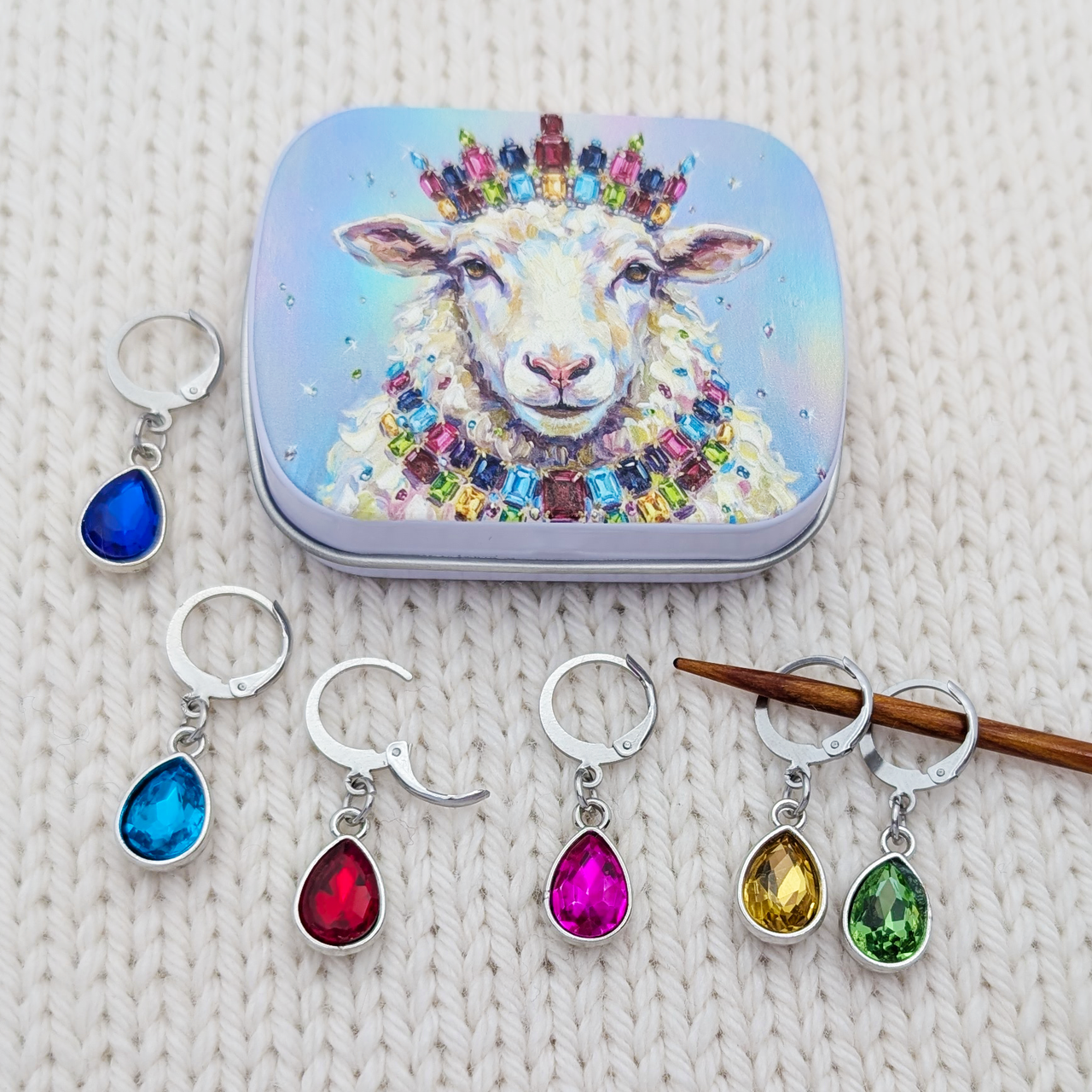 A set of silver teardrop-shaped locking stitch markers with colorful gemstones, displayed alongside a matching decorative tin featuring a sheep illustration. The charms are shown in multiple colors (blue, red, pink, yellow, green) on a textured cream knit background.