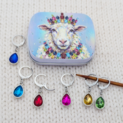 A set of silver teardrop-shaped locking stitch markers with colorful gemstones, displayed alongside a matching decorative tin featuring a sheep illustration. The charms are shown in multiple colors (blue, red, pink, yellow, green) on a textured cream knit background.