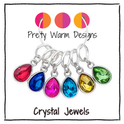 Six silver-tone ring style stitch markers with teardrop-shaped crystal glass gemstones in red, blue, pink, yellow, green, and light blue, displayed on a white background with 'Pretty Warm Designs' and 'Crystal Jewels' text.