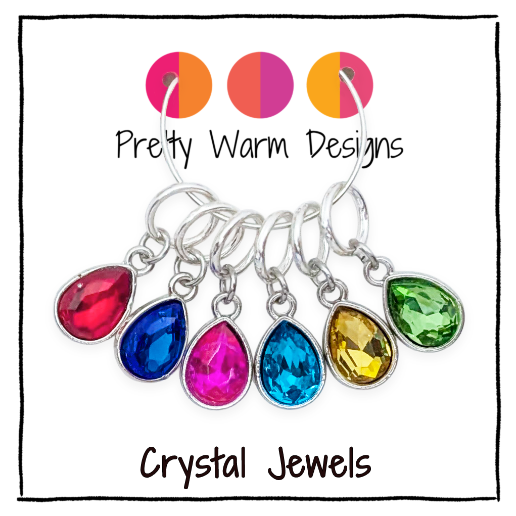 Six silver-tone ring style stitch markers with teardrop-shaped crystal glass gemstones in red, blue, pink, yellow, green, and light blue, displayed on a white background with 'Pretty Warm Designs' and 'Crystal Jewels' text.