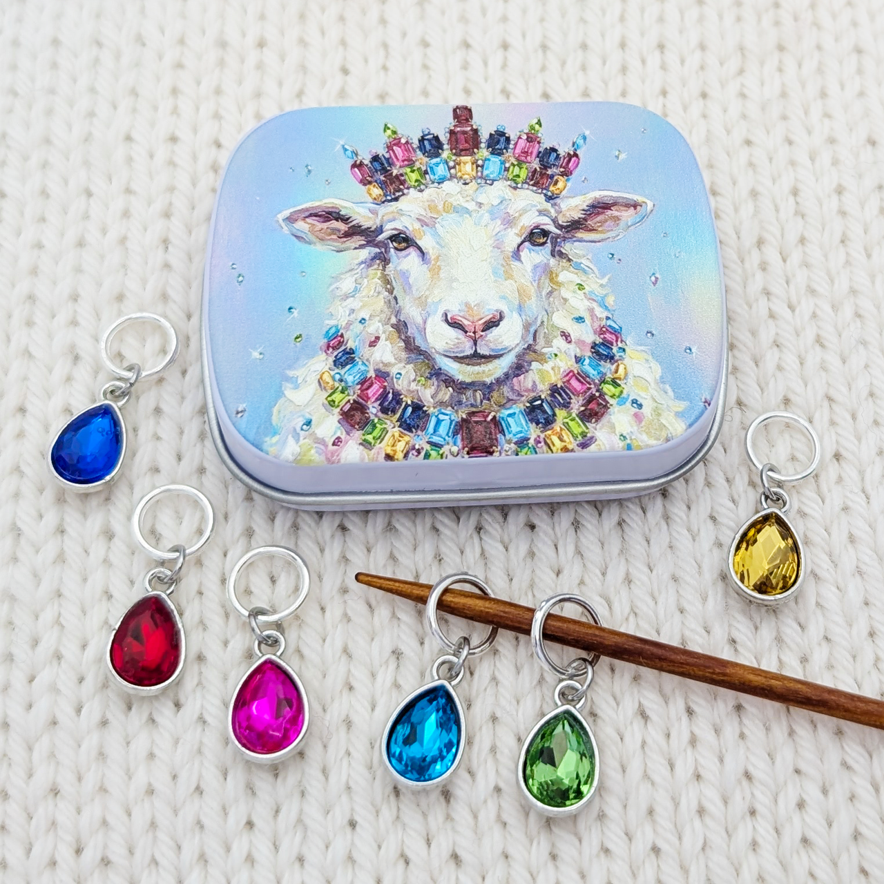 A set of colorful teardrop-shaped crystal glass stitch markers with silver rings, displayed alongside a small hinged tin featuring a jeweled sheep illustration. The markers are shown in various colors (blue, red, pink, green, yellow) on a white knitted fabric background.