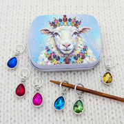 A set of colorful teardrop-shaped crystal glass stitch markers with silver rings, displayed alongside a small hinged tin featuring a jeweled sheep illustration. The markers are shown in various colors (blue, red, pink, green, yellow) on a white knitted fabric background.