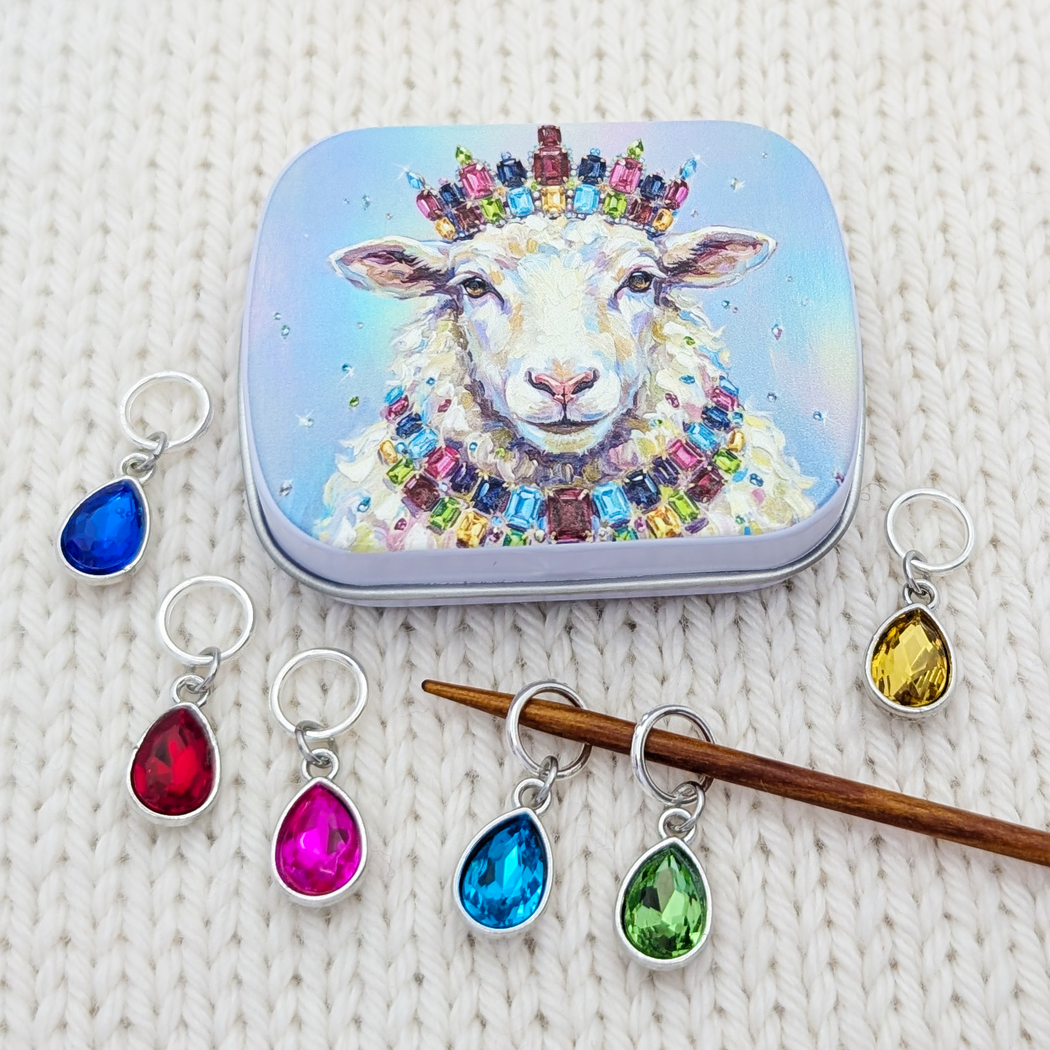 A set of colorful teardrop-shaped crystal glass stitch markers with silver rings, displayed alongside a small hinged tin featuring a jeweled sheep illustration. The markers are shown in various colors (blue, red, pink, green, yellow) on a white knitted fabric background.