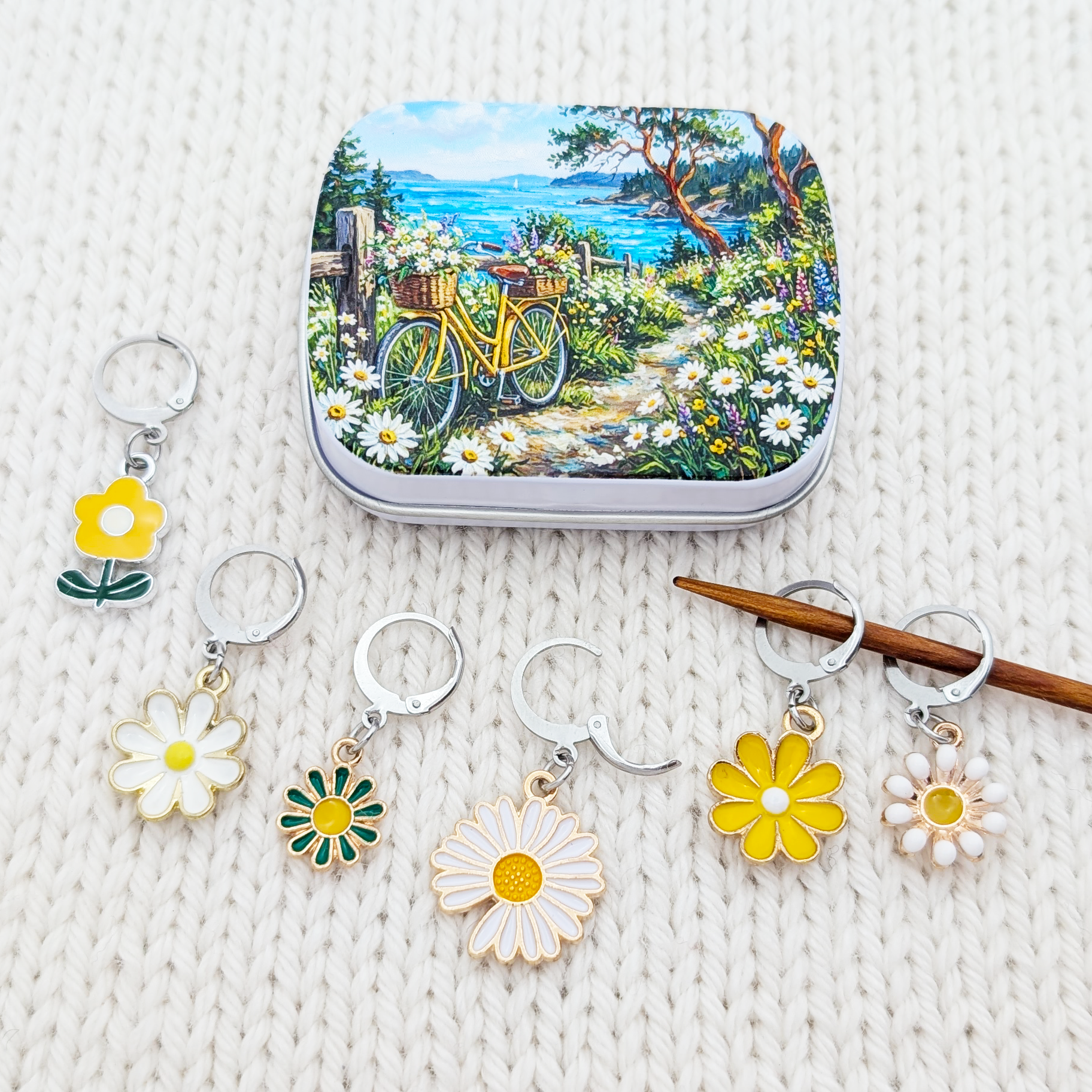 A set of flower-shaped knitting and crochet stitch holders and a matching small notions tin with a coastal garden illustration, arranged on a cream knit blanket.
