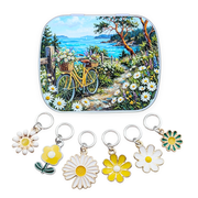 A hinged metal notions tin featuring a colorful coastal garden scene with a yellow bicycle, trees, flowers, and blue water, shown above five matching daisy flower-shaped charms with silver rings on a white background.