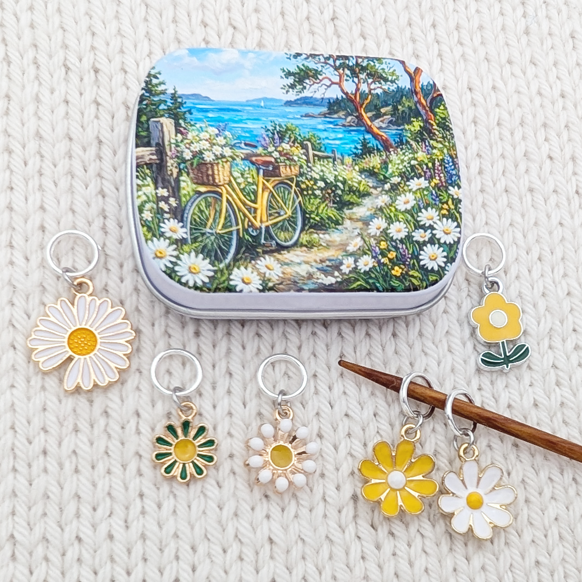 A decorative notions tin with a colorful coastal garden illustration sits on a cream knit blanket, surrounded by several daisy themed ring stitch markers and a knitting needle holding two more daisy ring markers.