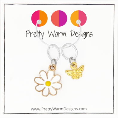 Two summer-themed knitting ring stitch markers, one yellow and white enamel daisy and one gold toned honeybee with Pretty Warm Designs text and logo