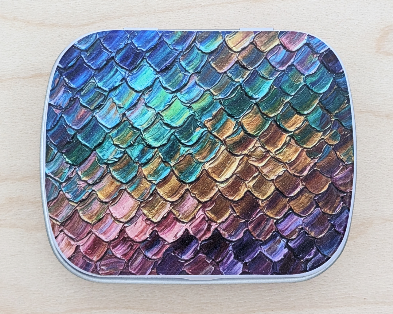 A hinged metal notions tin featuring a metallic iridescent dragon / mermaid scale pattern in shades of blue, green, gold, bronze, pink, and purple, set against a light wood background.
