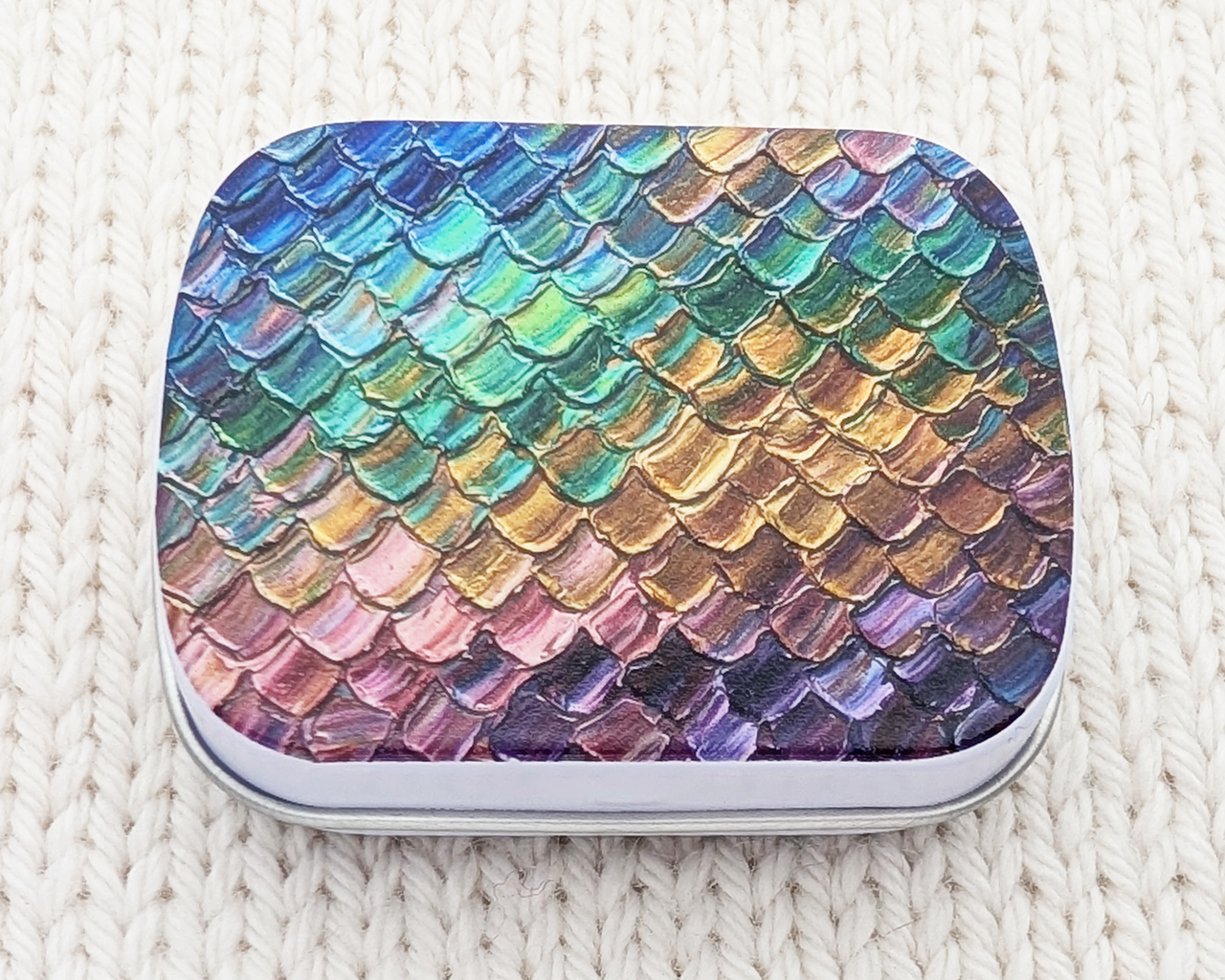 A hinged metal notions tin featuring a colorful iridescent dragon / mermaid scale pattern on the lid, resting on a cream-colored knitted blanket.