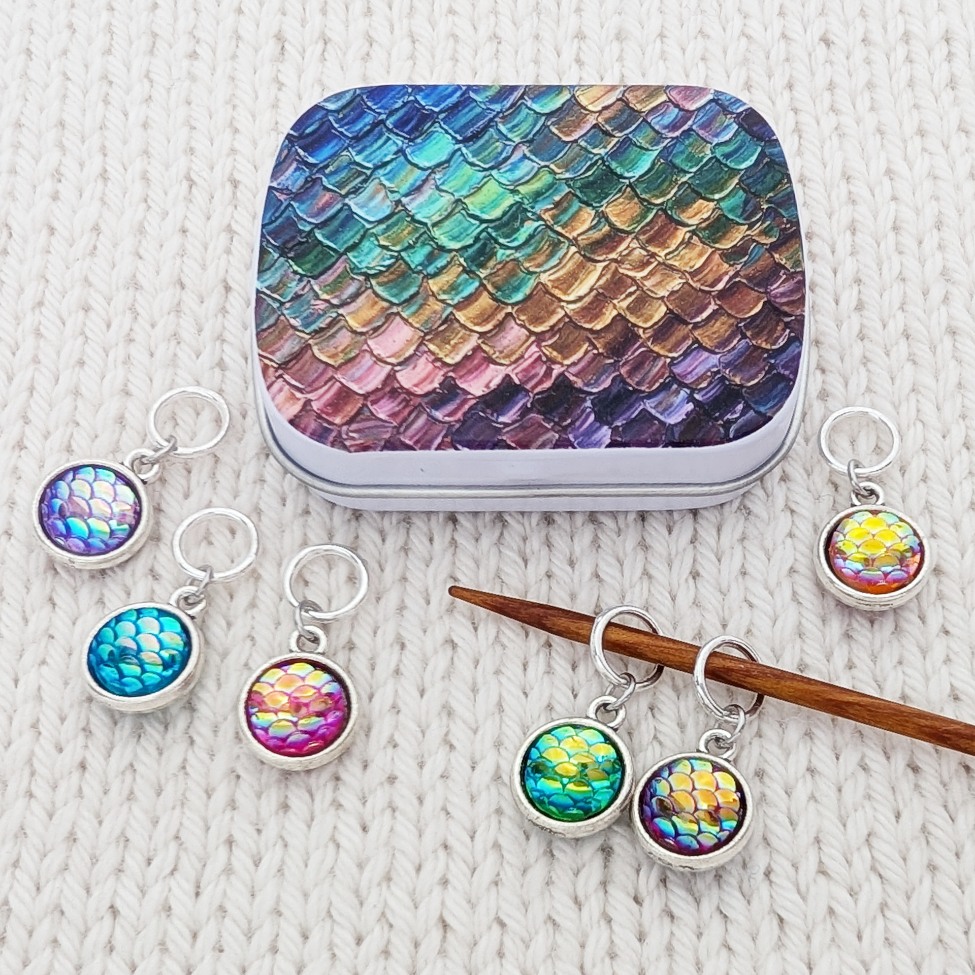 Dragon scale pattern knitting accessories displayed on a cream knit blanket, including a small iridescent metal notions tin and several silver jump rings with resin charms showing blue, green, purple, and pink scale designs, shown on a wooden knitting needle.