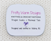 The bottom of a hinged metal notions tin with printed text reading 'Pretty Warm Designs, KNITTING & CROCHET NOTIONS, Dragon Scales + Mermaid Tails, Designed and crafted in Victoria, BC' and a small purple dragon graphic.