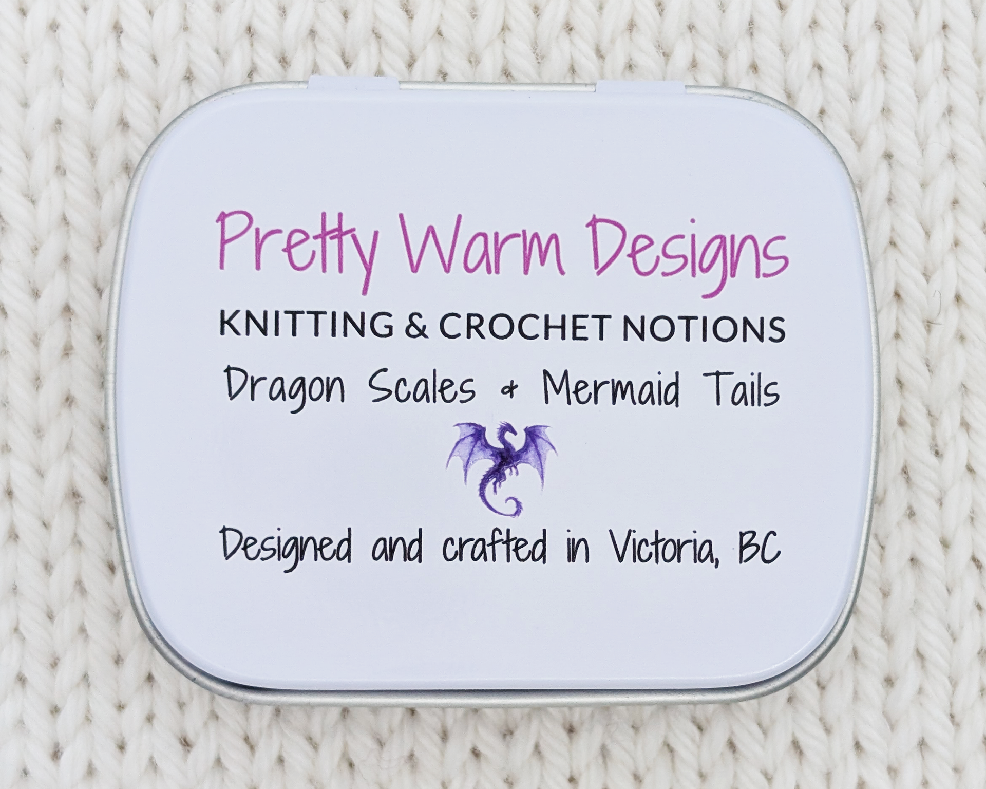 The bottom of a hinged metal notions tin with printed text reading 'Pretty Warm Designs, KNITTING & CROCHET NOTIONS, Dragon Scales + Mermaid Tails, Designed and crafted in Victoria, BC' and a small purple dragon graphic.