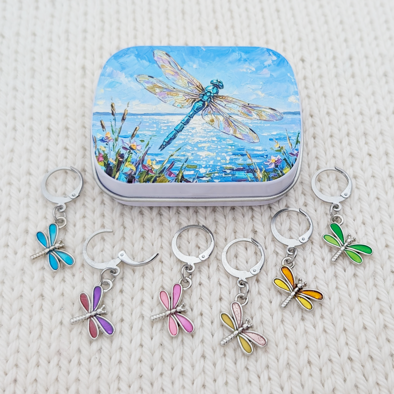 A set of five silver metal dragonfly-shaped crochet and knitting stitch markers with colorful enamel wings, displayed below their matching square tin. The tin features a blue dragonfly painting over water, all arranged on a cream-colored textured knit blanket.