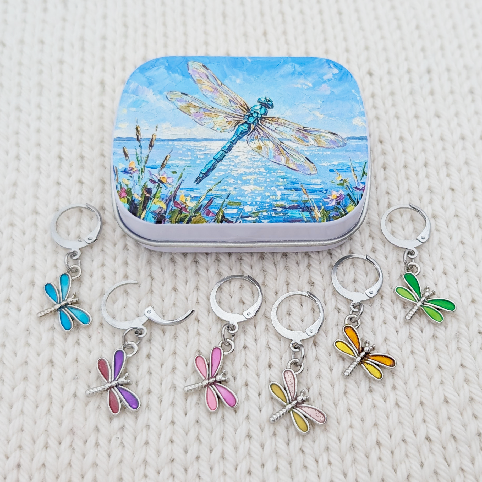 A set of five silver metal dragonfly-shaped crochet and knitting stitch markers with colorful enamel wings, displayed below their matching square tin. The tin features a blue dragonfly painting over water, all arranged on a cream-colored textured knit blanket.