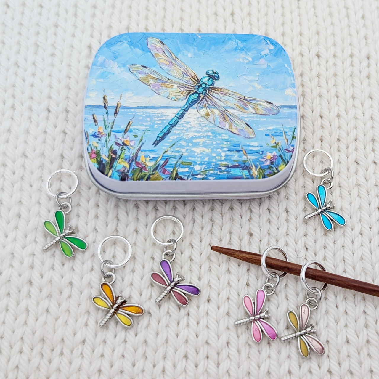 A tin of dragonfly-shaped knitting stitch markers arranged on a cream knit blanket, with several stitch markers in different colors shown alongside the tin, with a dragonfly artwork hinged storage tin.