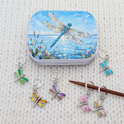 A tin of dragonfly-shaped knitting stitch markers arranged on a cream knit blanket, with several stitch markers in different colors shown alongside the tin, with a dragonfly artwork hinged storage tin.