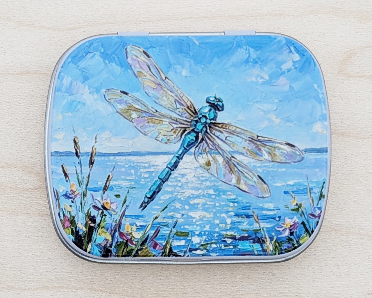 A hinged metal notions tin featuring a painted illustration of a blue dragonfly over water with wildflowers under a blue sky, placed on a light wood surface.