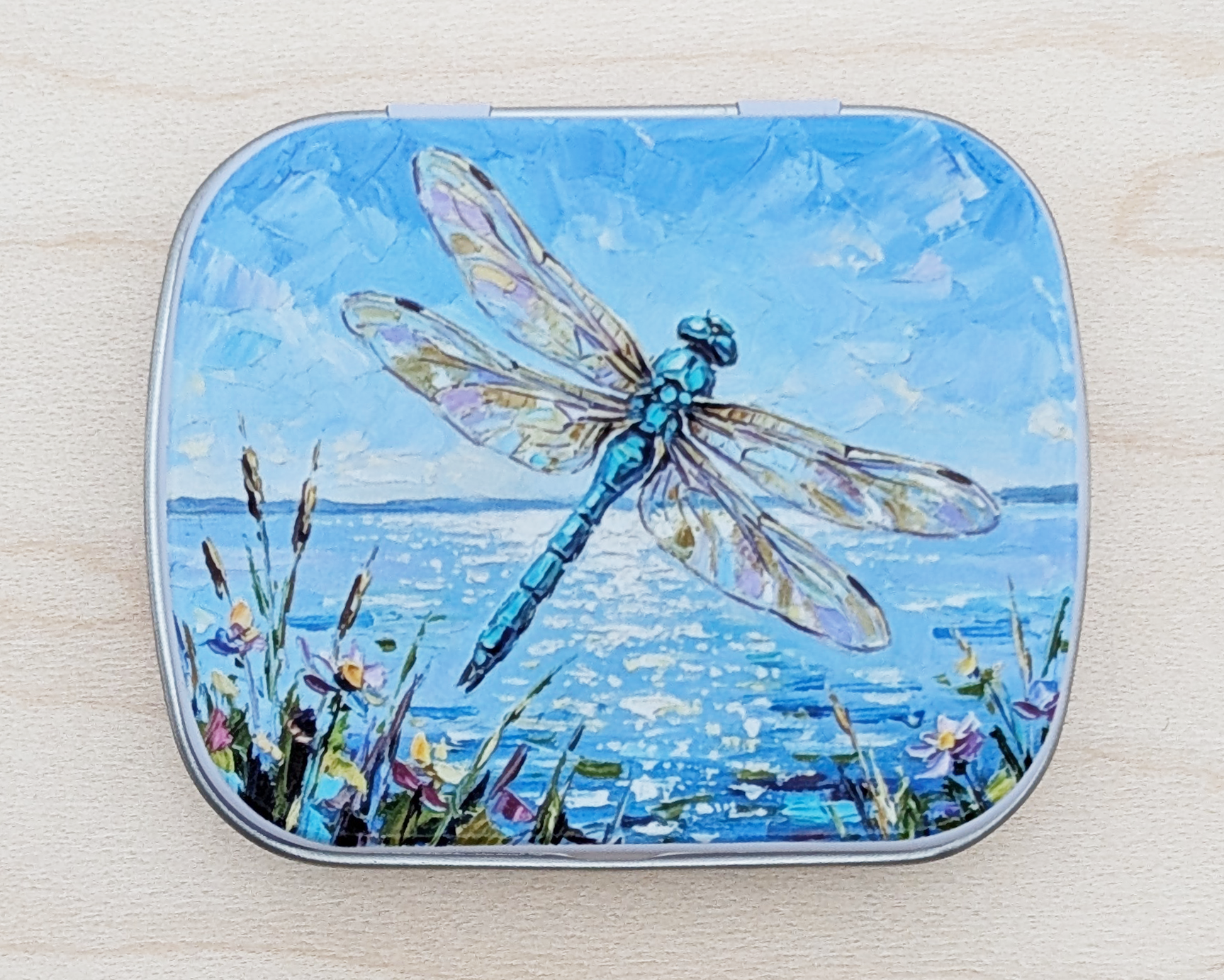 A hinged metal notions tin featuring a painted illustration of a blue dragonfly over water with wildflowers under a blue sky, placed on a light wood surface.