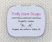 The bottom side of a hinged metal stitch marker tin printed with the text 'Pretty Warm Designs, KNITTING & CROCHET NOTIONS, Dragonfly Summer, Designed and crafted in Victoria, BC' along with two small dragonfly illustrations. The tin is photographed on a cream-colored knitted fabric background.
