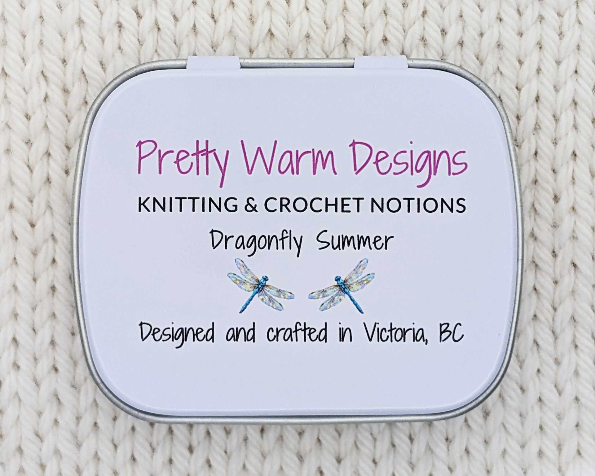 The bottom side of a hinged metal stitch marker tin printed with the text 'Pretty Warm Designs, KNITTING & CROCHET NOTIONS, Dragonfly Summer, Designed and crafted in Victoria, BC' along with two small dragonfly illustrations. The tin is photographed on a cream-colored knitted fabric background.
