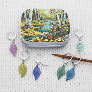 A small craft storage tin with an autumn forest scene with birch trees, a stream, and a squirrel, shown above six locking crochet stitch markers with nature themed lightweight filigree metal charms