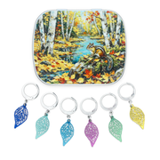 A crochet notions tin with an autumn forest scene with birch trees, a stream, and a squirrel, shown above six locking stitch markers with lightweight filigree metal charms in the shape of leaves.