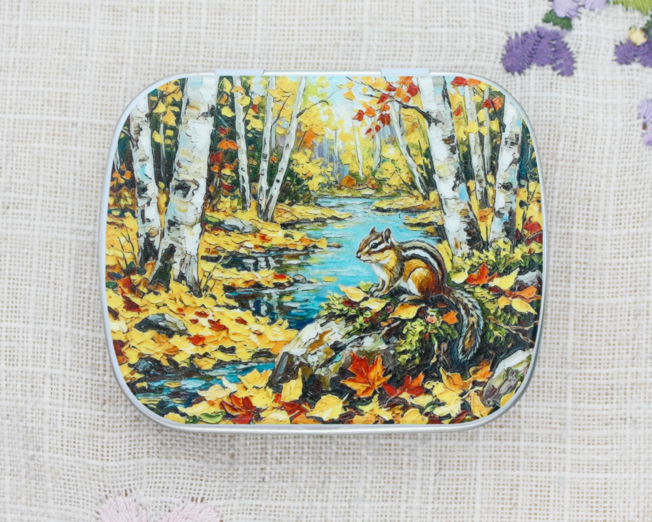 A hinged metal notions tin featuring a Canadian scene with a chipmunk amongst the autumn leaves, beside a freshwater stream.