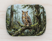 A hinged metal notions tin featuring a detailed illustration of a great horned owl perched on a mossy tree branch in a forest. The tin is shown on a light wood surface.