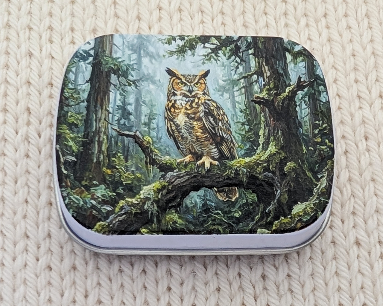 A hinged metal notions tin featuring a detailed illustration of a great horned owl perched on a mossy branch in a forest. The tin is placed on a cream-colored, textured knit blanket.