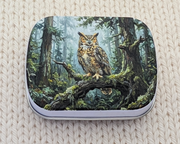 A hinged metal notions tin featuring a detailed illustration of a great horned owl perched on a mossy branch in a forest. The tin is placed on a cream-colored, textured knit blanket.