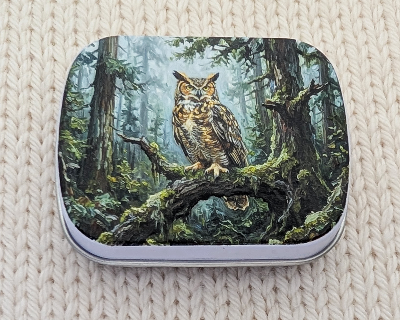 A hinged metal notions tin featuring a detailed illustration of a great horned owl perched on a mossy branch in a forest. The tin is placed on a cream-colored, textured knit blanket.