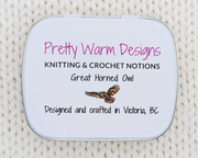 The bottom of a hinged metal notions tin with the text 'Pretty Warm Designs, KNITTING & CROCHET NOTIONS, Great Horned Owl, Designed and crafted in Victoria, BC' and an owl illustration, placed on a cream-colored knitted fabric background.