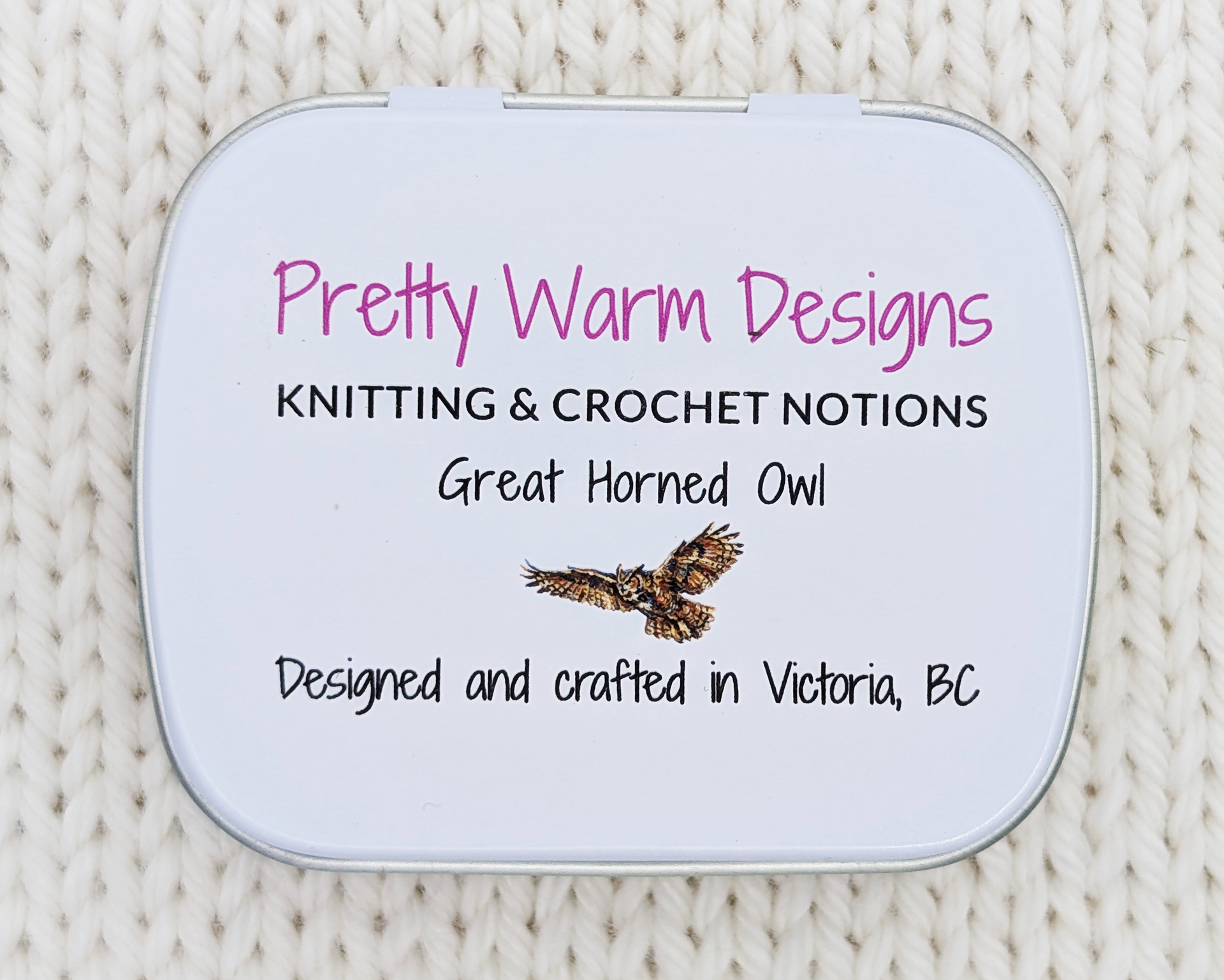 The bottom of a hinged metal notions tin with the text 'Pretty Warm Designs, KNITTING & CROCHET NOTIONS, Great Horned Owl, Designed and crafted in Victoria, BC' and an owl illustration, placed on a cream-colored knitted fabric background.