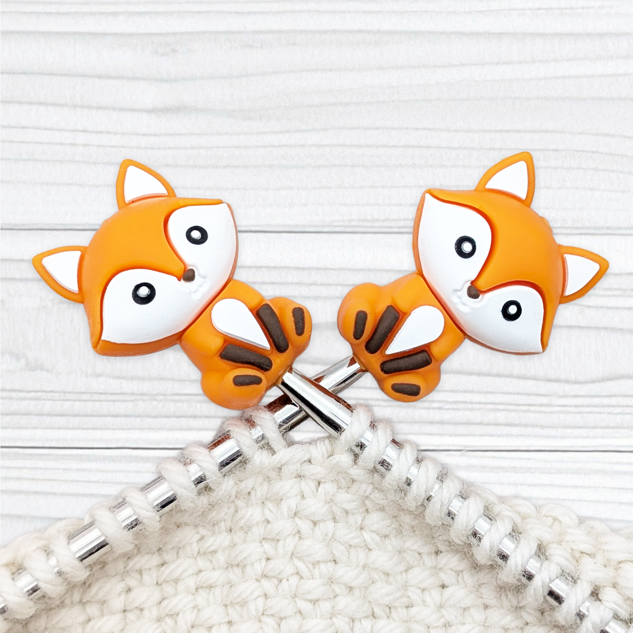 Pair of Crafty Fox stitch stoppers on knitting needle tips, showing orange fox faces with white cheeks and ears