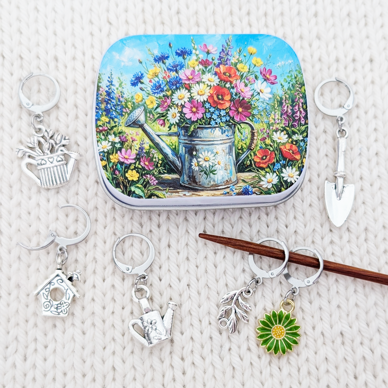 A square tin with a colorful floral and watering can illustration is arranged on a white knit surface with several silver progress keepers including a birdhouse, watering can, trowel, flower, and leaf designs.