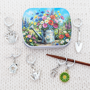 A square tin with a colorful floral and watering can illustration is arranged on a white knit surface with several silver progress keepers including a birdhouse, watering can, trowel, flower, and leaf designs.