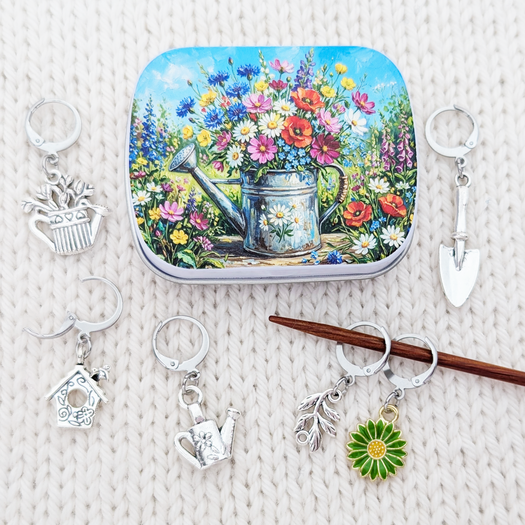 A square tin with a colorful floral and watering can illustration is arranged on a white knit surface with several silver progress keepers including a birdhouse, watering can, trowel, flower, and leaf designs.