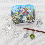 A square tin with rounded corners featuring a colorful painting of a watering can filled with wildflowers, arranged on a cream knit fabric background with several small silver and ring stitch markers for knitters around it.