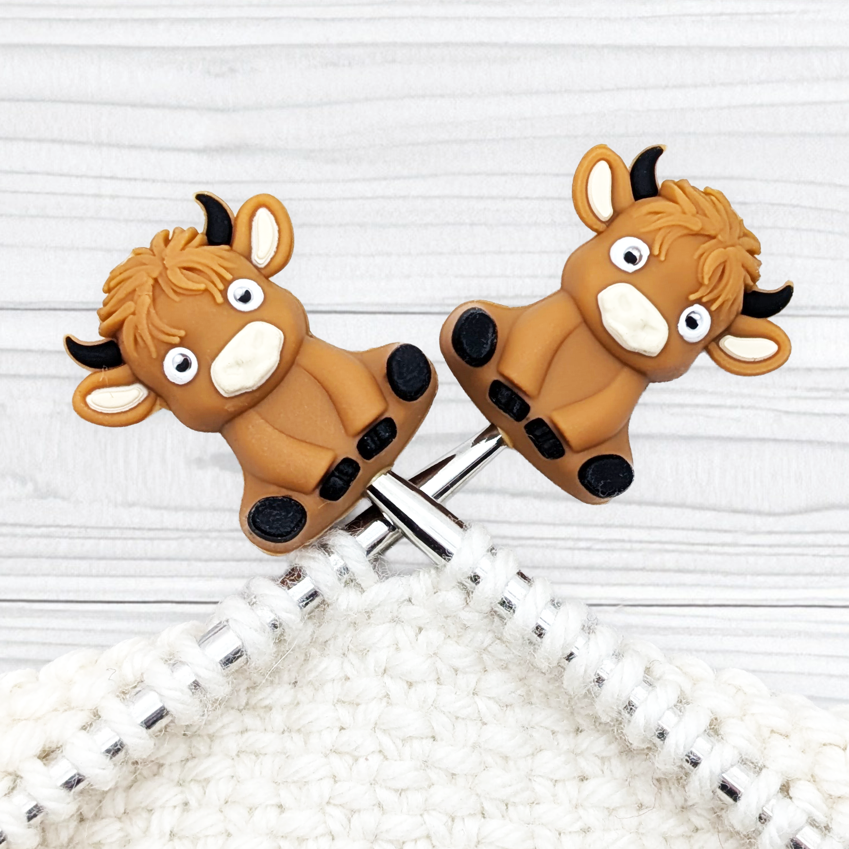 Pair of Och Aye the Moo Highland Cow stitch stoppers sitting on knitting needle tips, showing shaggy fringe and cheeky faces