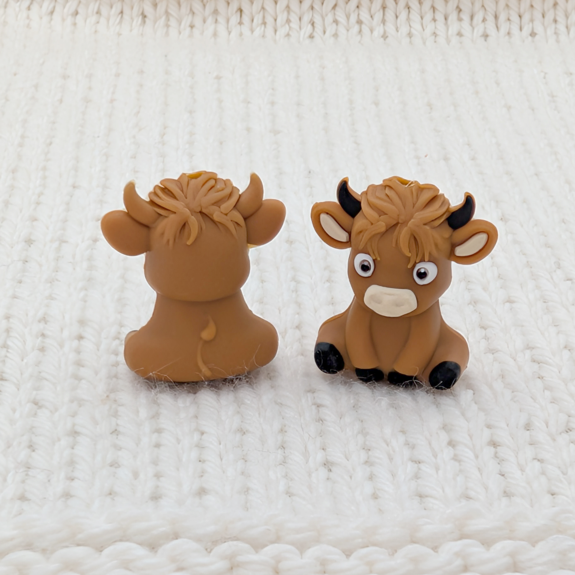 Front and back view of Highland Cow stitch stoppers on white knit fabric, showing sculpted 3D detail including textured fringe, black horns and hooves.