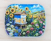 A hinged metal notions tin featuring a colorful painted illustration of a beehive surrounded by sunflowers, wildflowers, and a blue sky, placed on a light wood surface.