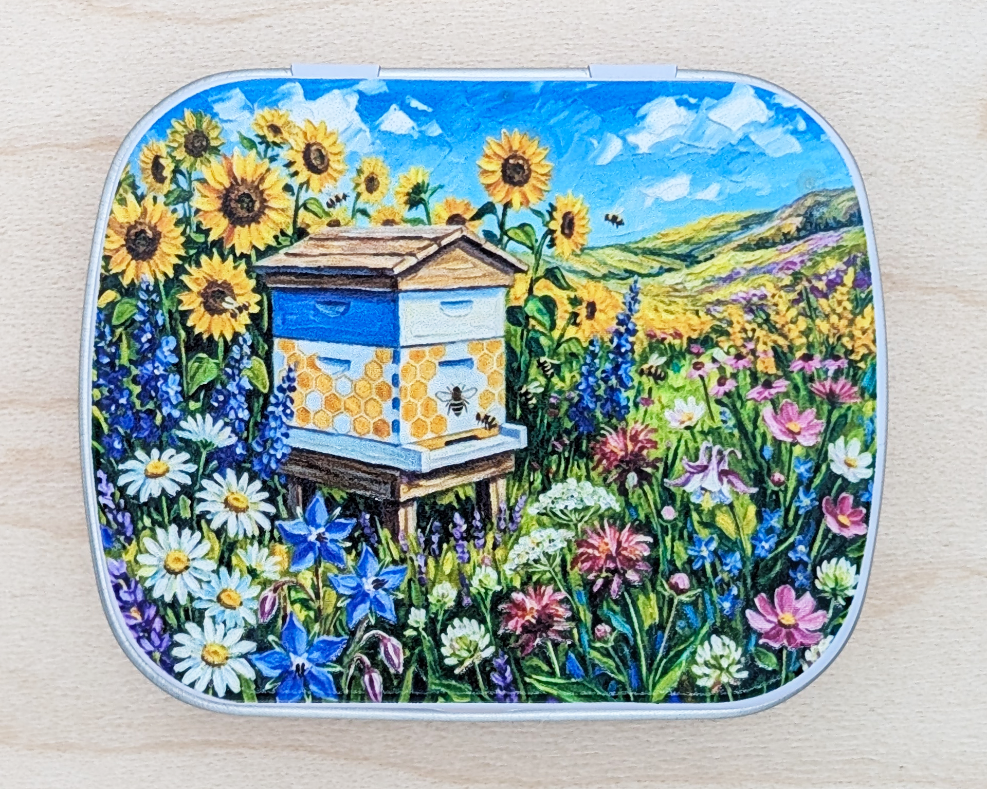 A hinged metal notions tin featuring a colorful painted illustration of a beehive surrounded by sunflowers, wildflowers, and a blue sky, placed on a light wood surface.
