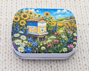 A hinged metal notions tin featuring a colorful printed design of a beehive surrounded by sunflowers, bluebells, and wildflowers under a blue sky. The tin is placed on a white textured knit blanket.
