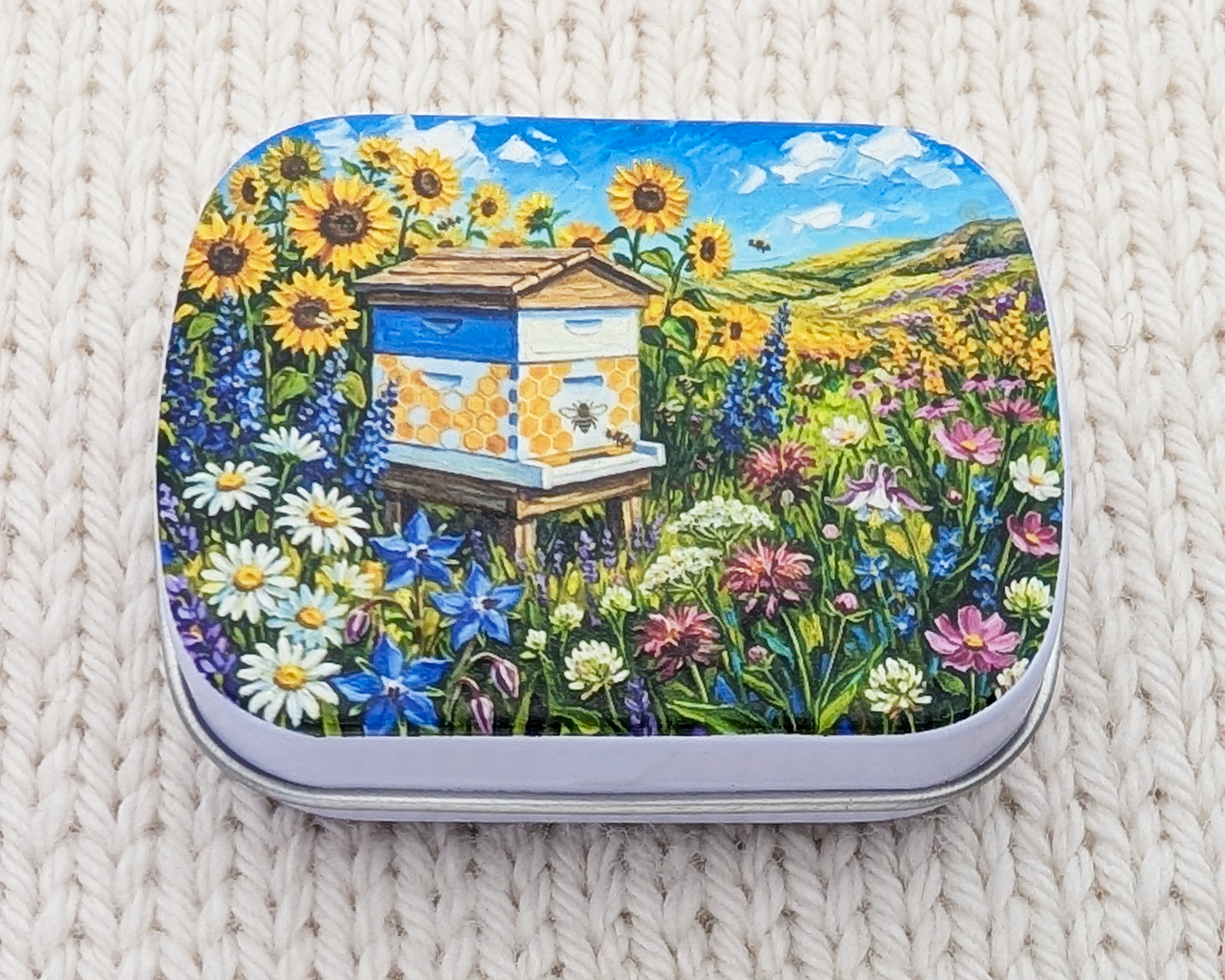A hinged metal notions tin featuring a colorful printed design of a beehive surrounded by sunflowers, bluebells, and wildflowers under a blue sky. The tin is placed on a white textured knit blanket.