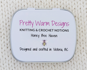 The bottom of a metal notions tin with printed text that reads 'Pretty Warm Designs', 'KNITTING & CROCHET NOTIONS', 'Honey Bee Haven', and 'Designed and crafted in Victoria, BC' with a small bee illustration. The tin is photographed from above on a cream-colored knitted fabric background.