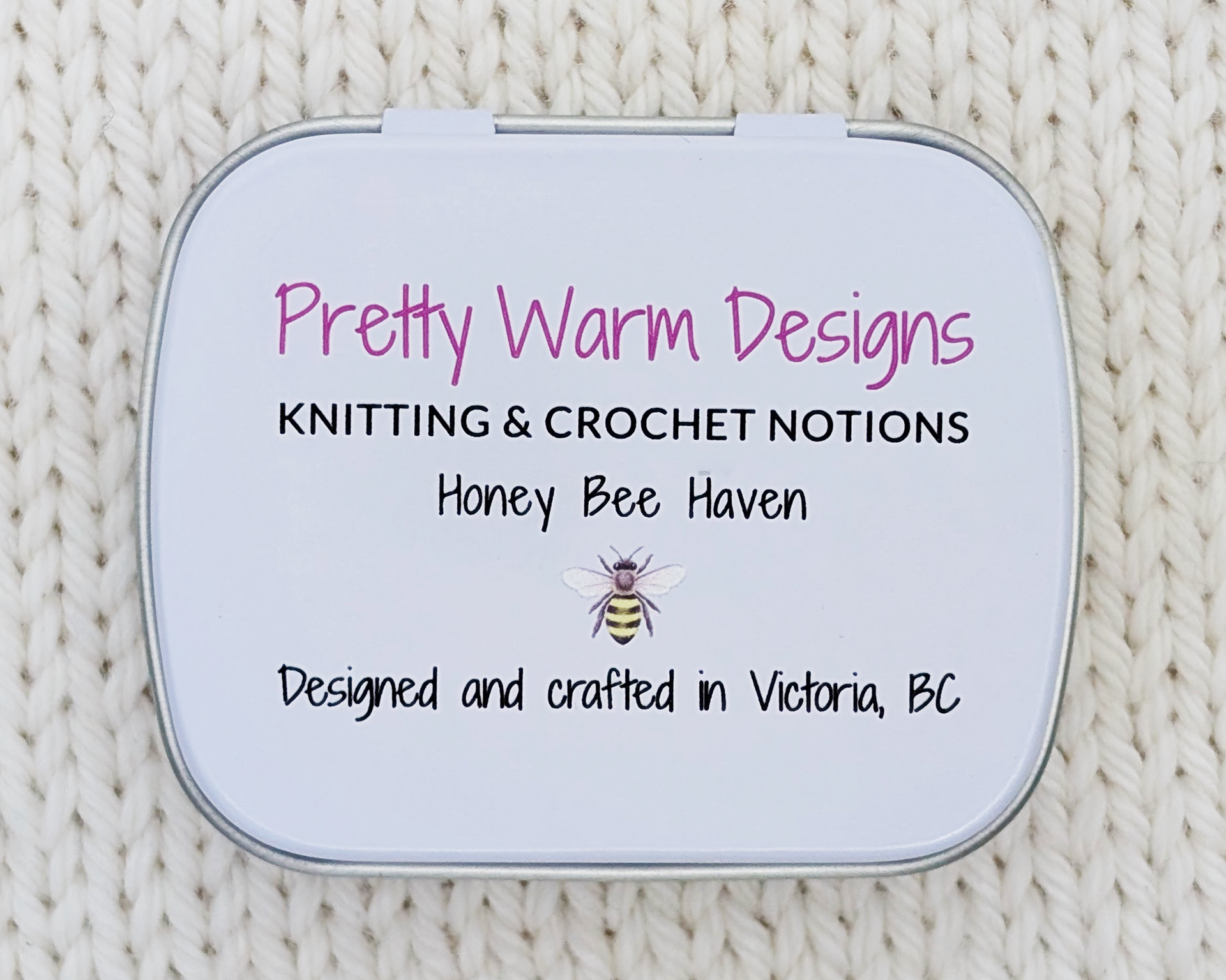 The bottom of a metal notions tin with printed text that reads 'Pretty Warm Designs', 'KNITTING & CROCHET NOTIONS', 'Honey Bee Haven', and 'Designed and crafted in Victoria, BC' with a small bee illustration. The tin is photographed from above on a cream-colored knitted fabric background.