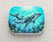 A hinged metal notions tin for knitting and crochet accessories featuring a humpback whale mother and calf