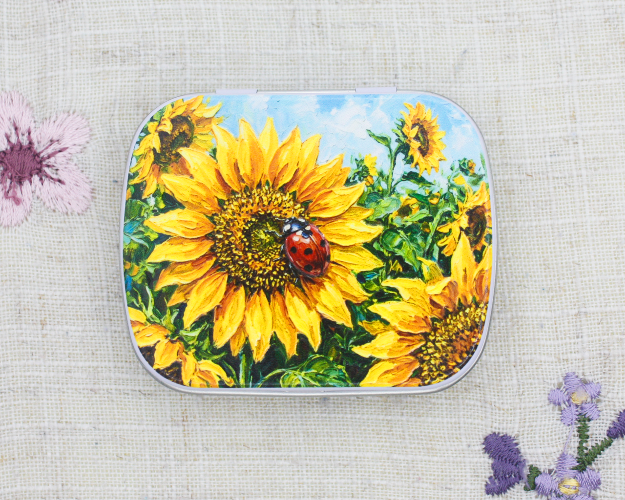 A metal notions tin featuring a vibrant print of yellow sunflowers and a red ladybug on the lid, set against a textured beige linen background with embroidered flowers.