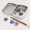 Set of six enamel butterfly charm snag free ring stitch markers in red, yellow, green, blue, pink and purple on knitting needle resting on white knitted square with storage tin for knitting by Pretty Warm Designs