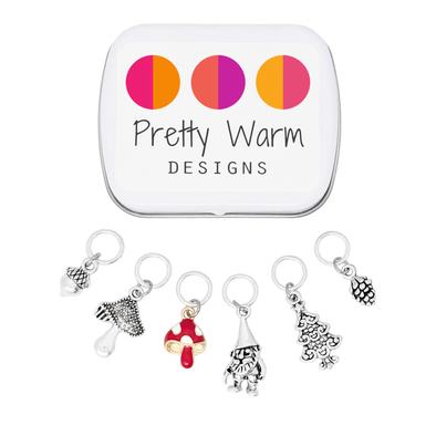 Set of six stitch markers for knitting, including a red enamel toadstool, as well as an antique silver acorn, toadstool, woodland gnome, pine cone and Christmas tree with decorative storage tin labeled with Pretty Warm Designs text and logo sold by Pretty Warm Designs Inc.